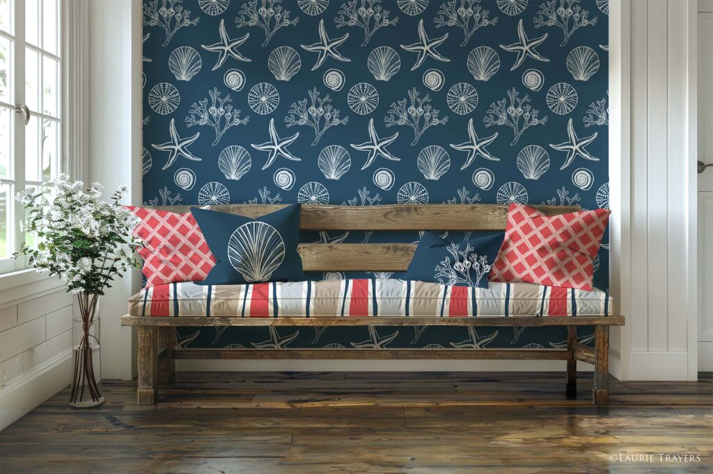 A coastal‑style entryway featuring dark blue wallpaper with white hand‑drawn seashells, seaweed, and starfish. A reclaimed‑wood bench holds seat cushions in a wavy stripe fabric of dark blue, coral, and sand. Two coral basket‑pattern throw pillows sit on the far left and right, along with a dark blue square pillow with a white scallop shell and a rectangular dark blue pillow with white seaweed. Copyright: Laurie Trayers.