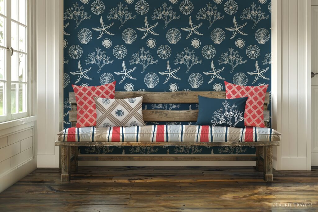 A coastal‑style entryway featuring dark blue wallpaper with white hand‑drawn seashells, seaweed, and starfish. A reclaimed‑wood bench holds seat cushions in a wavy stripe fabric of dark blue, coral, and sand. Two coral basket‑pattern throw pillows sit on the far left and right, along with a dark blue square pillow with a white scallop shell and a rectangular dark blue pillow with white seaweed. Copyright: Laurie Trayers.