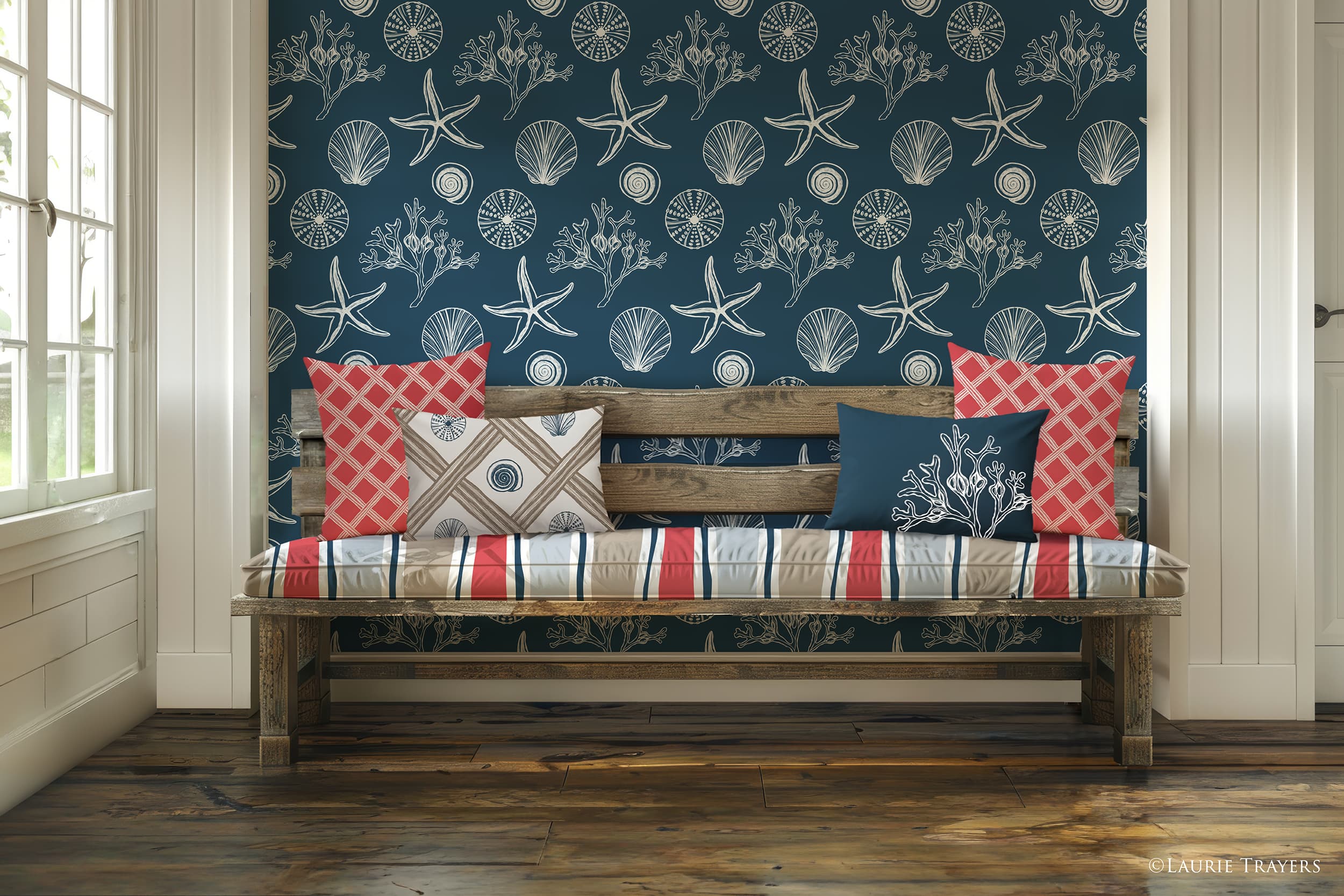 A coastal‑style entryway featuring dark blue wallpaper with white hand‑drawn seashells, seaweed, and starfish. A reclaimed‑wood bench holds seat cushions in a wavy stripe fabric of dark blue, coral, and sand. Two coral basket‑pattern throw pillows sit on the far left and right, along with a dark blue square pillow with a white scallop shell and a rectangular dark blue pillow with white seaweed. Copyright: Laurie Trayers.